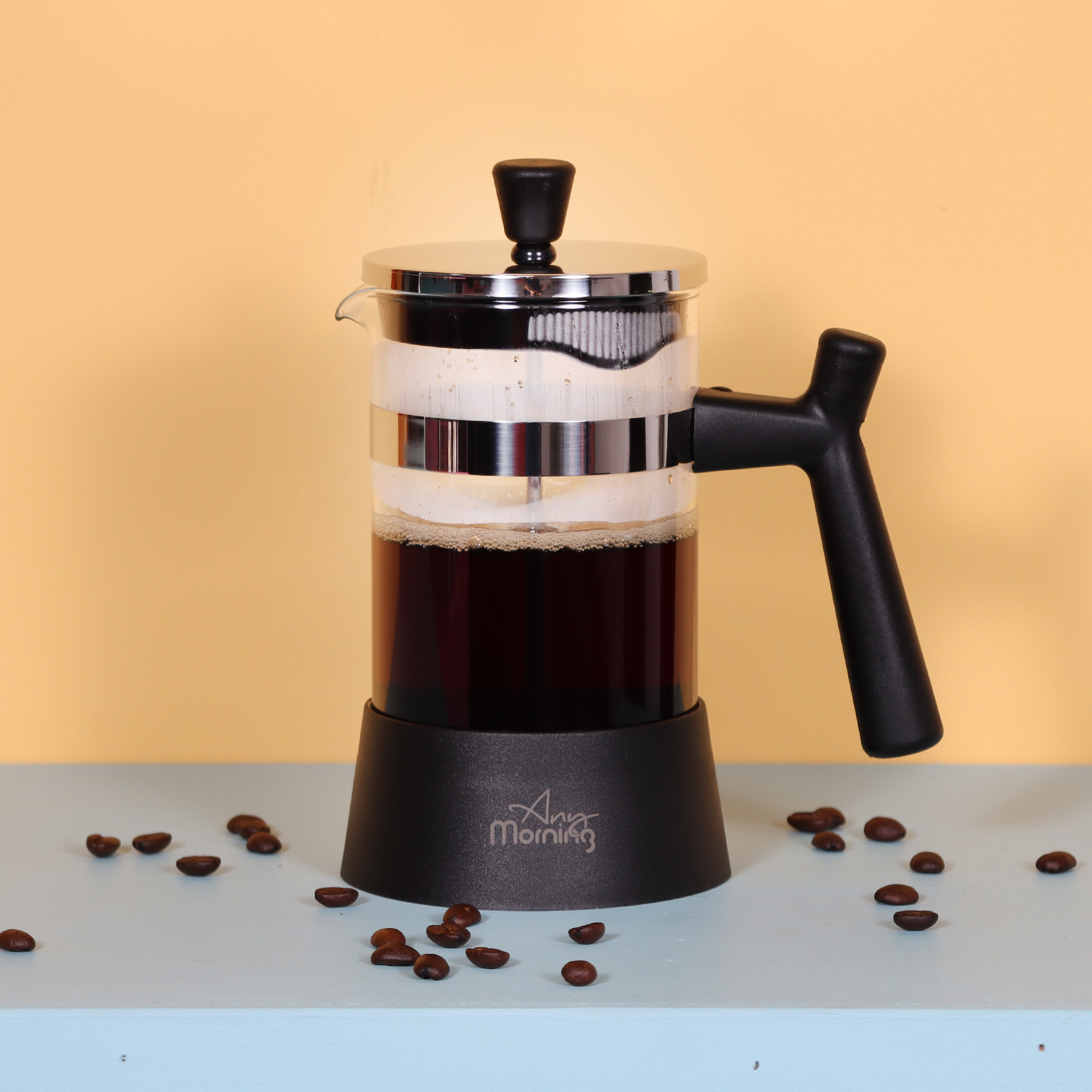 Una prensa francesa con café dentro, rodeada de granos de café sobre una superficie azul claro, fondo amarillo.