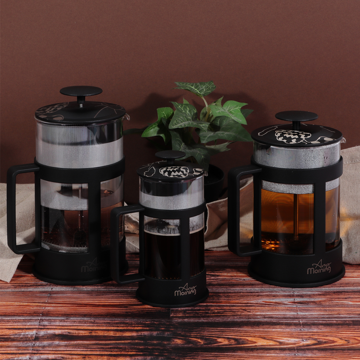 Tres prensas francesas negras con té sobre una superficie de madera. Una planta en el fondo. Paño de lino a ambos lados.