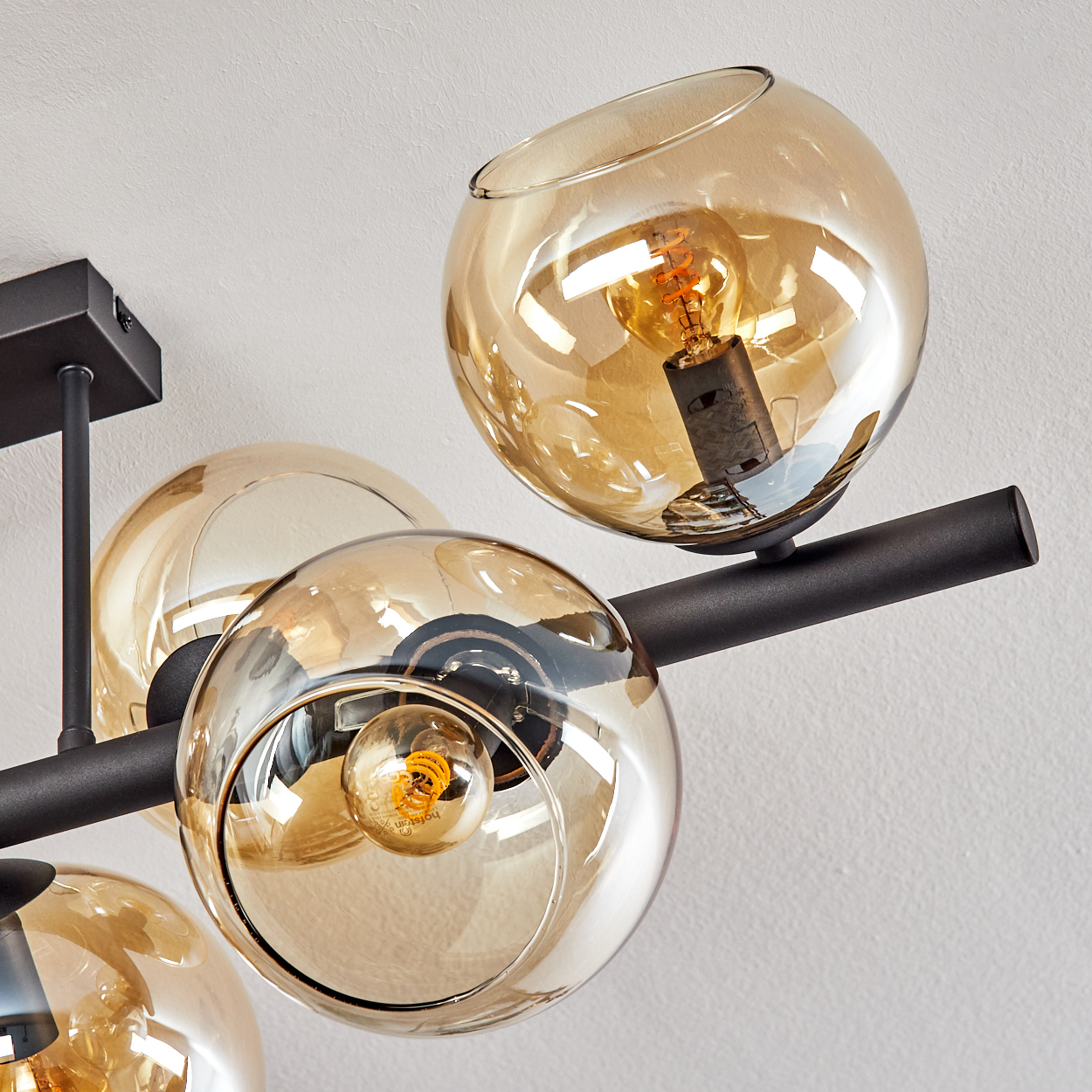 Eine moderne Deckenlampe mit vier bernsteinfarbenen Glaskugeln, in denen sich jeweils eine Glühbirne befindet, an einem schwarzen Metallrahmen an einer weißen Wand befestigt.