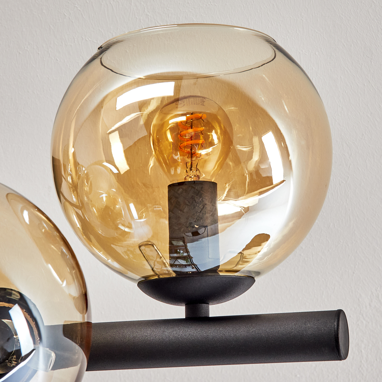 Nahaufnahme einer Leuchte. Ein kugelförmiger, bernsteinfarbener Glasschirm umschließt eine leuchtende Glühbirne. Schwarzer Metallarm und -sockel vor weißer Wand.