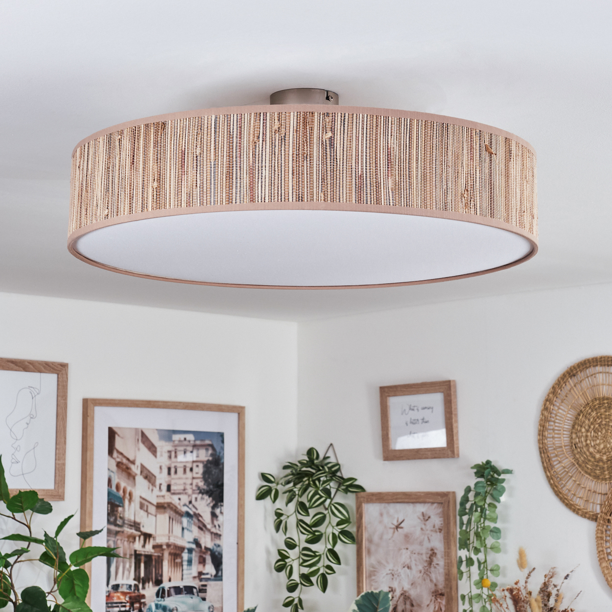 Eine runde Deckenlampe mit gewebtem Schirm. Darunter schmücken gerahmte Bilder und Pflanzen eine weiße Wand. Der Raum ist hell und einladend.
