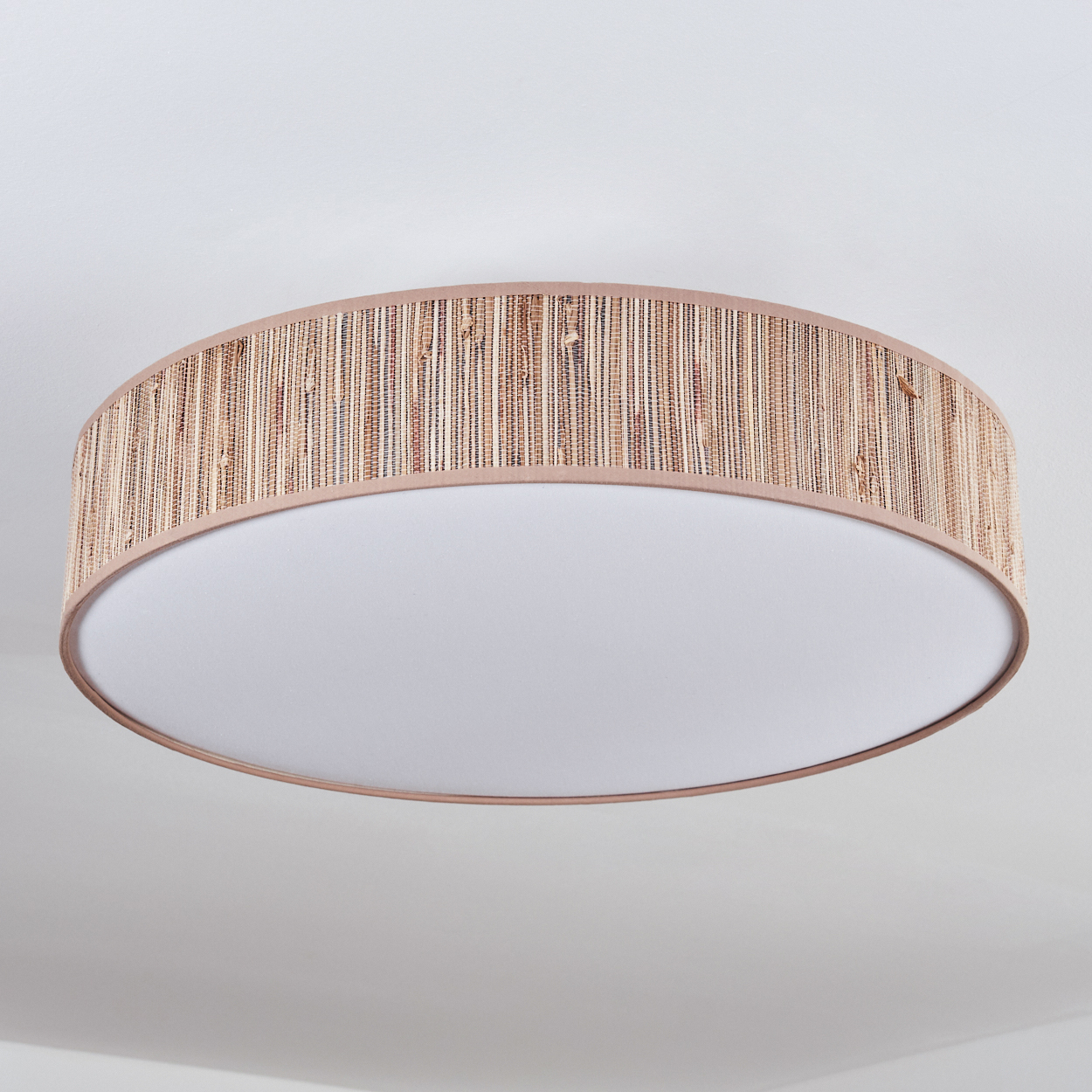 Eine runde Deckenleuchte mit einem gewebten, hellbraunen Schirm und einem weißen, kreisförmigen Lichtdiffusor. Der Hintergrund ist eine weiße Decke.