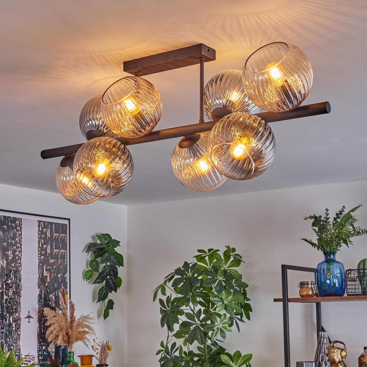 Eine moderne Deckenleuchte mit Glaskugeln, Pflanzen und einem Regal mit Dekorationen. Das Licht erhellt den Raum. Ein Bild hängt an der Wand.