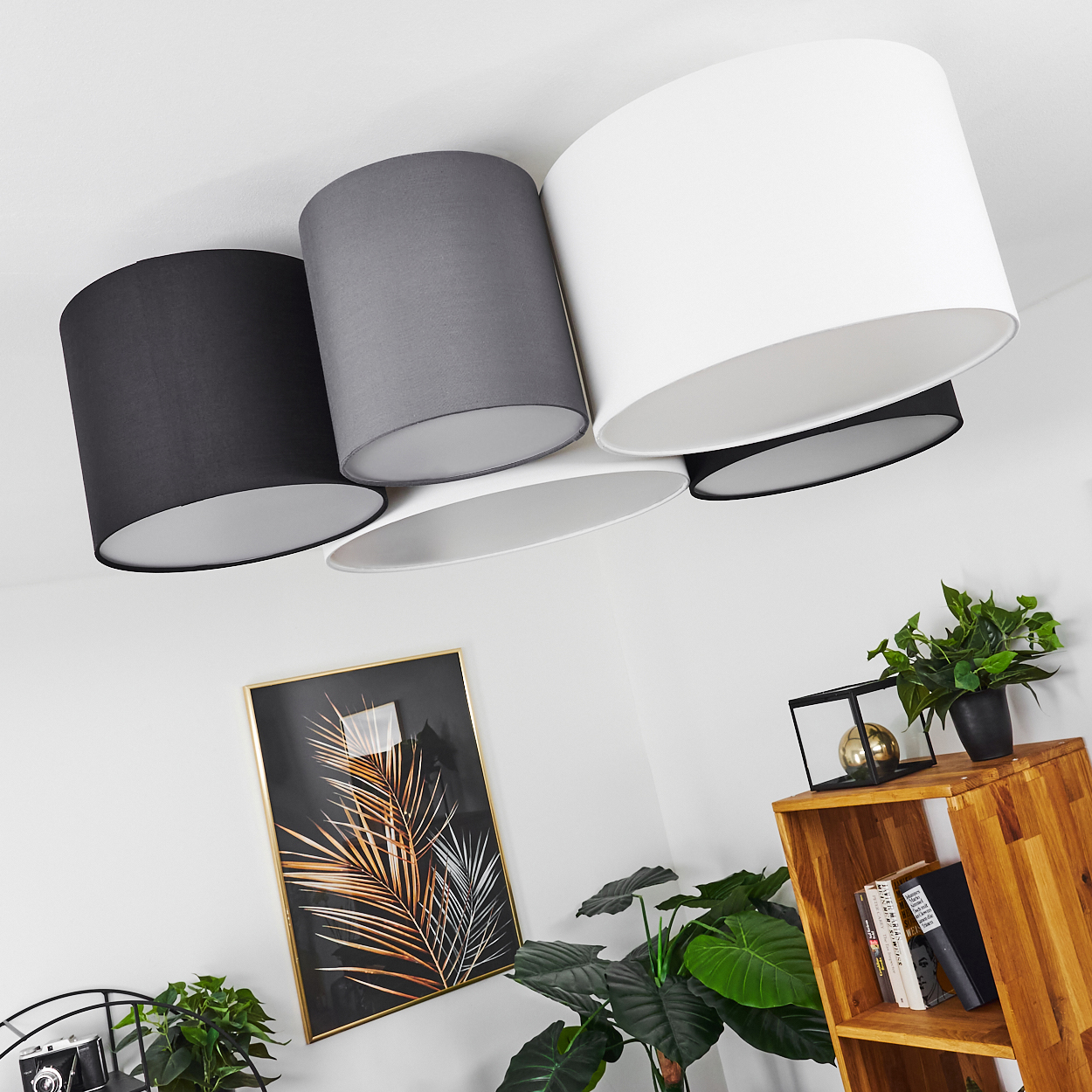 Vier Lampenschirme hängen von der Decke, schwarz, grau und weiß. Ein Bild mit goldenen Blättern hängt an der Wand, mit Pflanzen und einem Holzregal.
