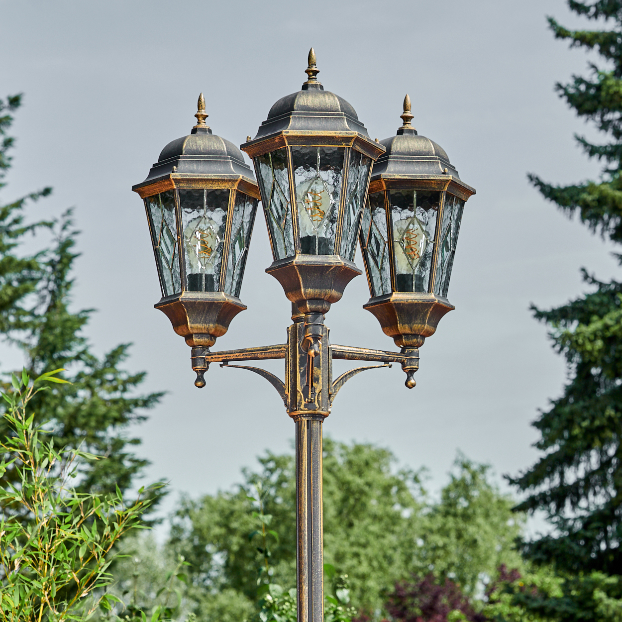 Eine verzierte bronzefarbene Straßenlaterne mit drei Lampen, grüne Bäume und Laub im Hintergrund, hellgrauer Himmel.
