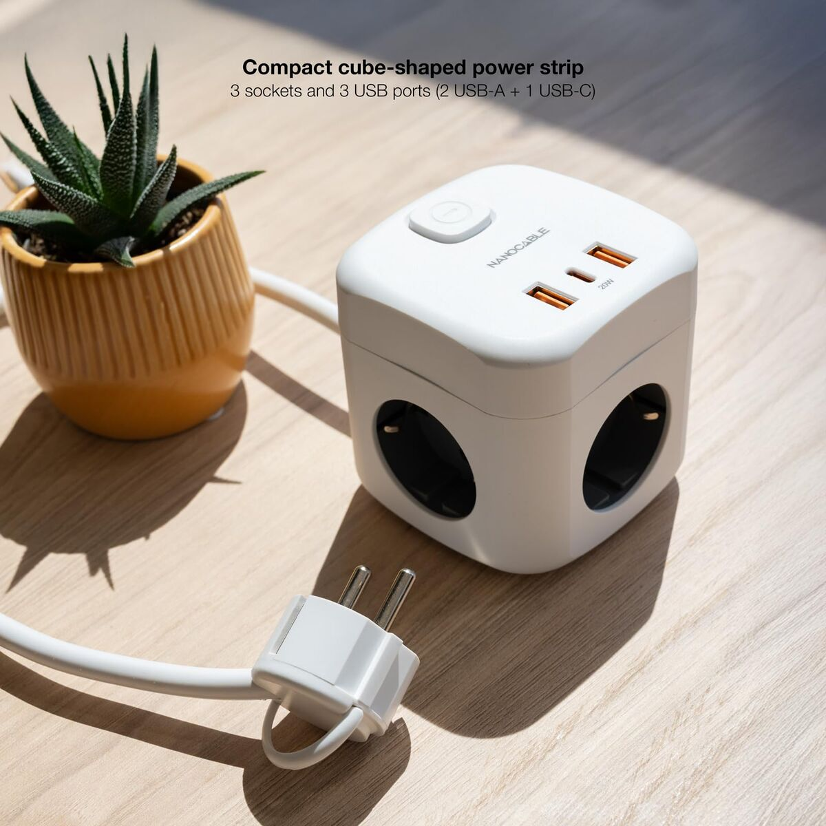 Ein weißer Würfel-Steckdosenleiste mit Steckdosen und USB-Anschlüssen. Daneben steht eine kleine Topfpflanze. Ein Stecker ist mit dem Würfel verbunden.