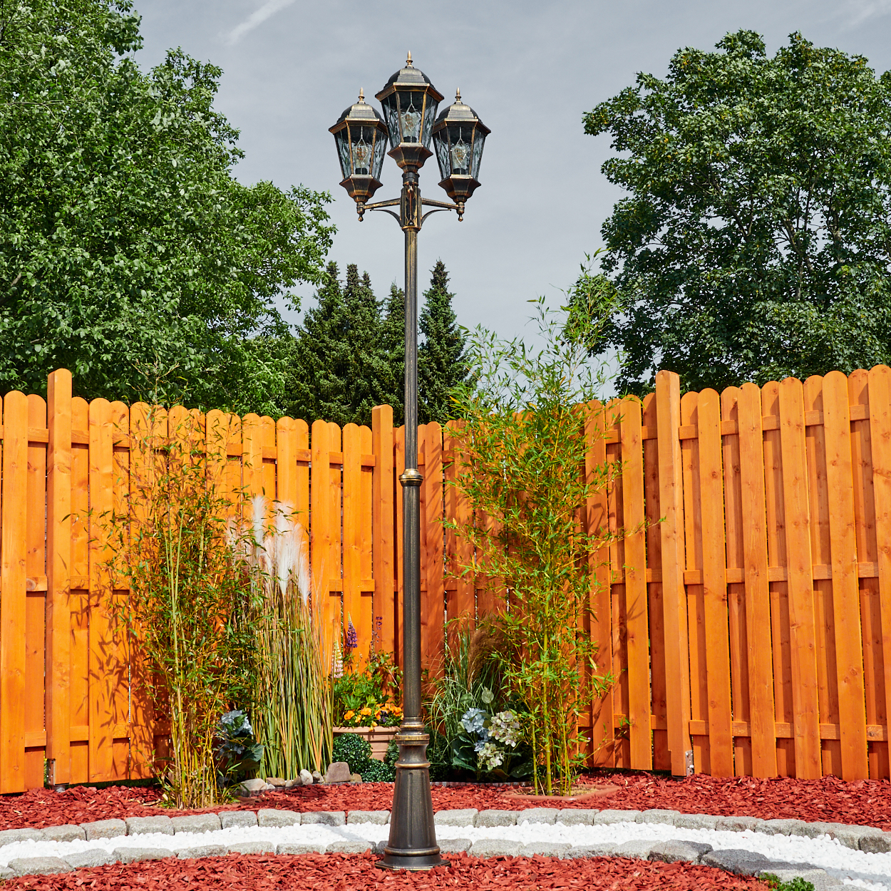 Eine verzierte Laterne in einem Garten, umgeben von einem orangefarbenen Zaun, Pflanzen und Bäumen unter einem bewölkten Himmel.