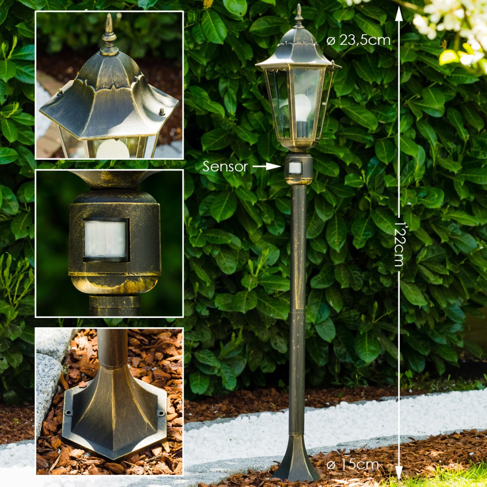 Eine bronzefarbene Außenlampe mit Sensor steht hoch. Sie ist 122 cm hoch, mit einem 23,5 cm breiten Oberteil und 15 cm breiten Sockel, vor grünem Laub.
