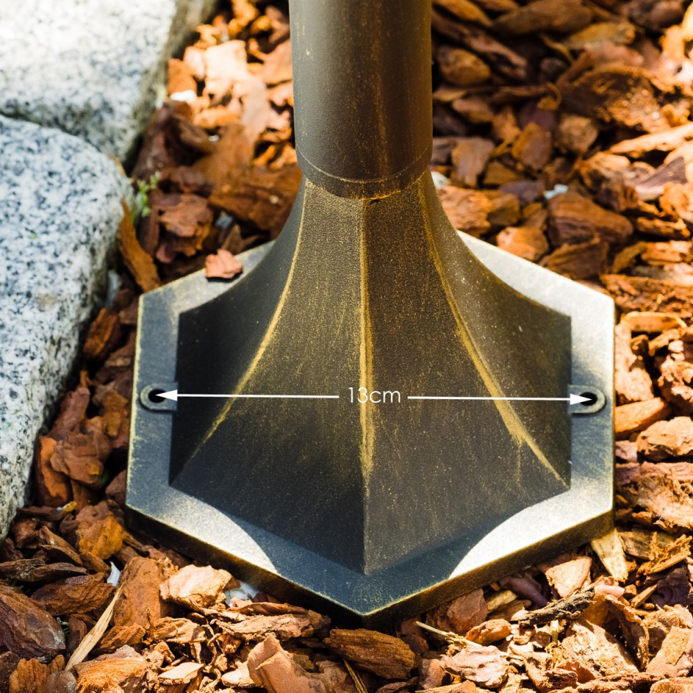Nahaufnahme zeigt Metallbasis einer Lampe. Sie ist schwarz mit goldenen Akzenten und steht auf braunem Mulch, mit 13 cm Messung.