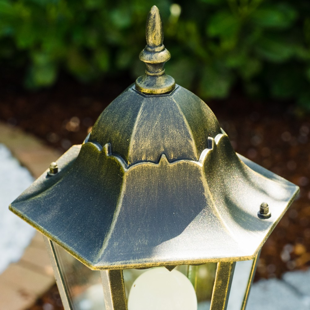 Nahaufnahme einer dekorativen Außenlampe mit schwarzem und goldenem Finish. Grünes Laub ist im Hintergrund. Im Inneren ist ein weißes Licht zu sehen.