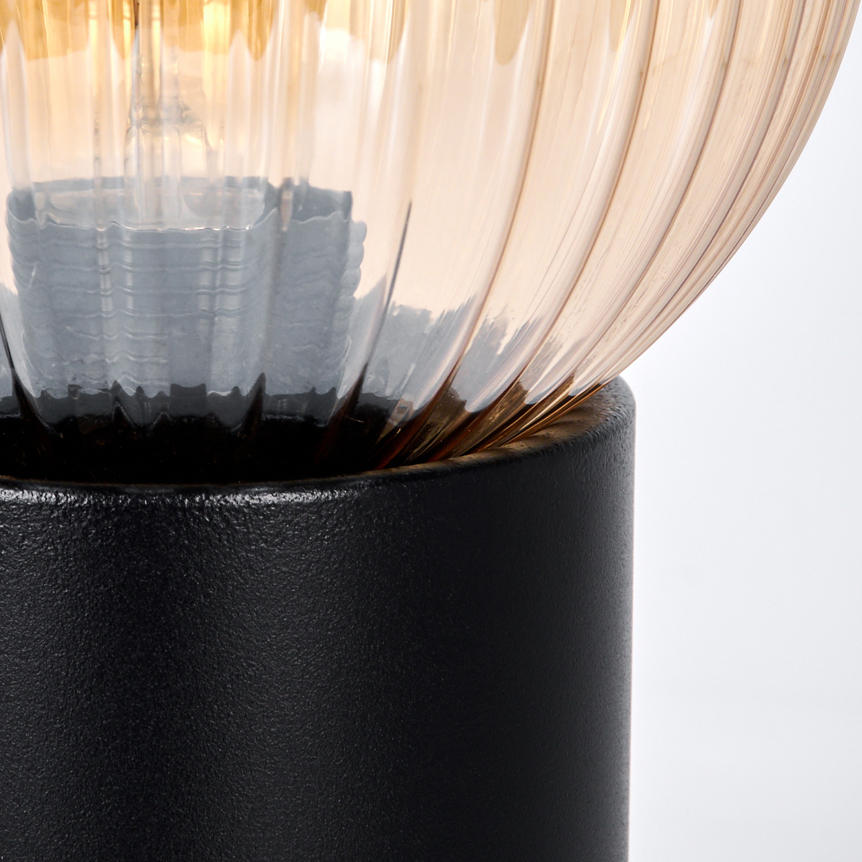 Nahaufnahme einer Lampe. Schwarzer Sockel, gerippter, bernsteinfarbener Glasschirm. Glühfaden der Glühbirne sichtbar.