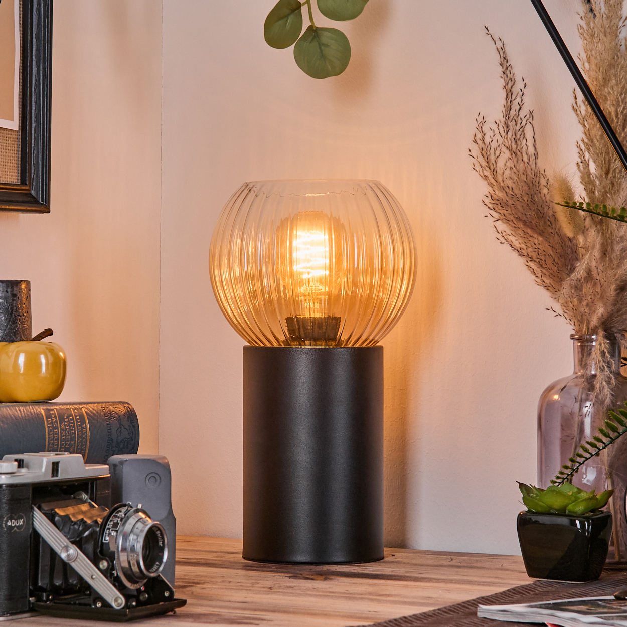 Eine leuchtende Lampe mit Glasschirm steht auf einer Holzoberfläche. Daneben Bücher, eine Kamera, Pflanzen und Dekoration.