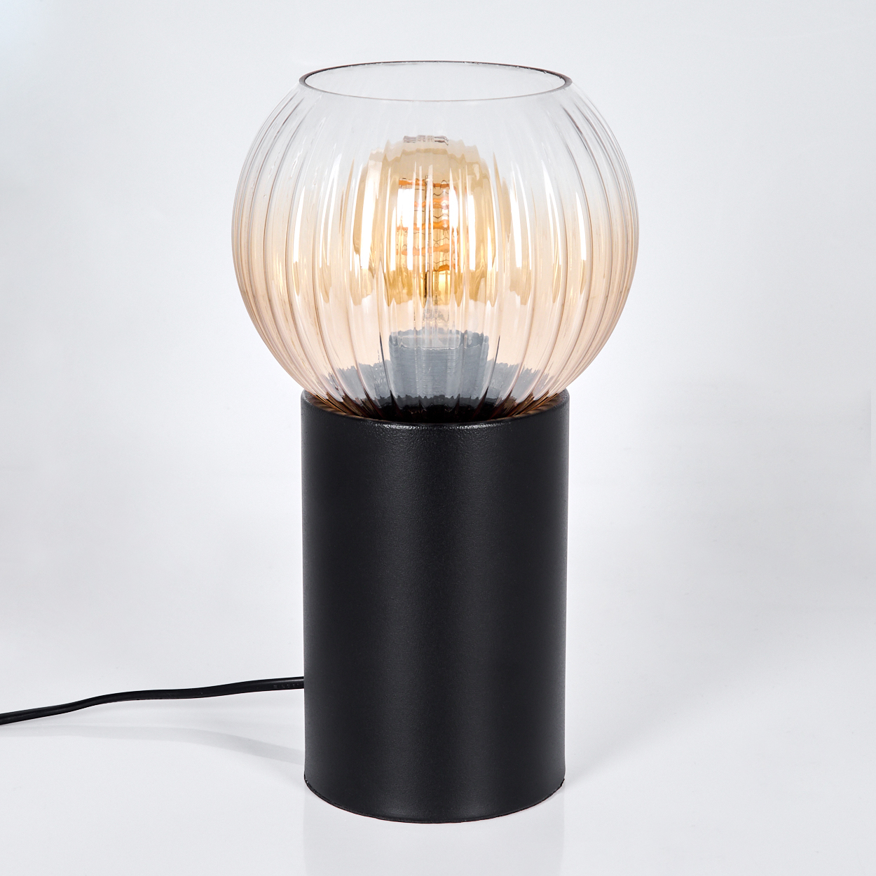 Eine moderne Lampe mit schwarzem zylindrischen Sockel. Sie hat einen kugelförmigen, gerippten Glasschirm mit warmem Orangeglühen.