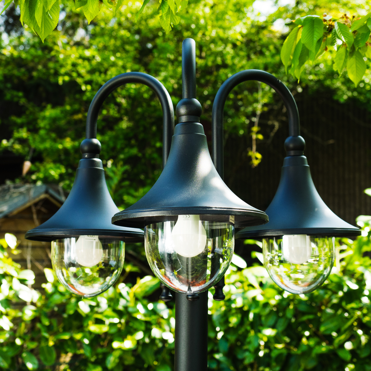 Schwarze Außenleuchte mit drei Lampen und Glaskugeln, beleuchtet. Grünes Laub im Hintergrund, mit Blättern im Vordergrund.