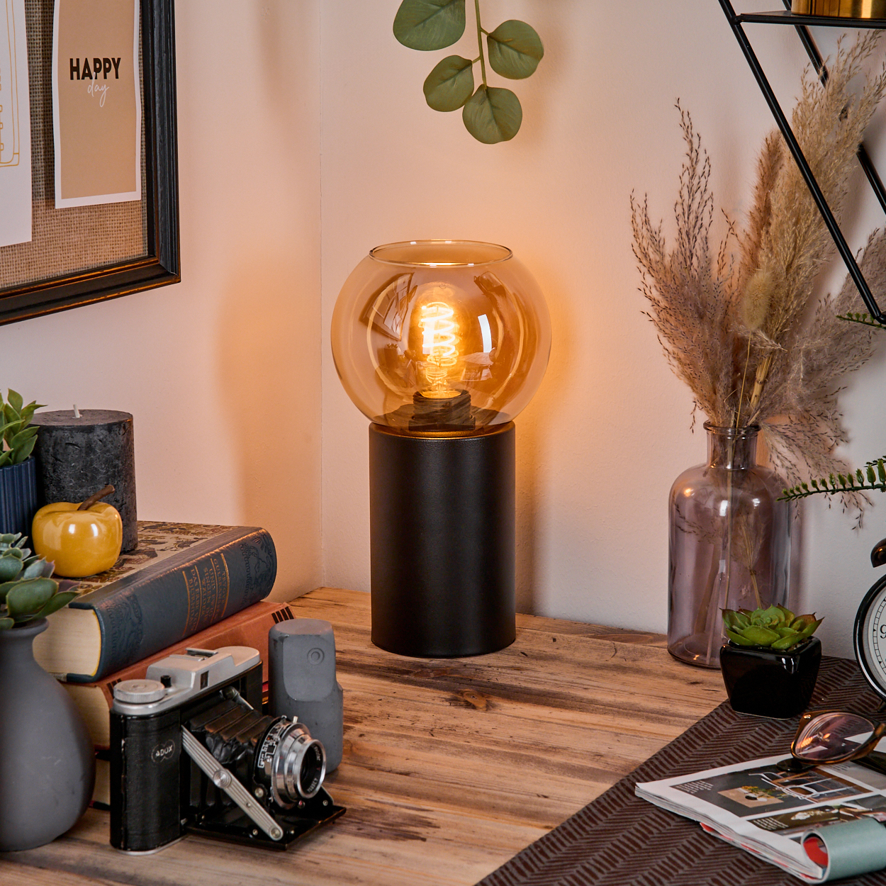 Eine brennende Lampe steht auf einem Holztisch mit Büchern, einer Kamera und Dekoration. Im Hintergrund steht eine Vase mit Pampasgras.