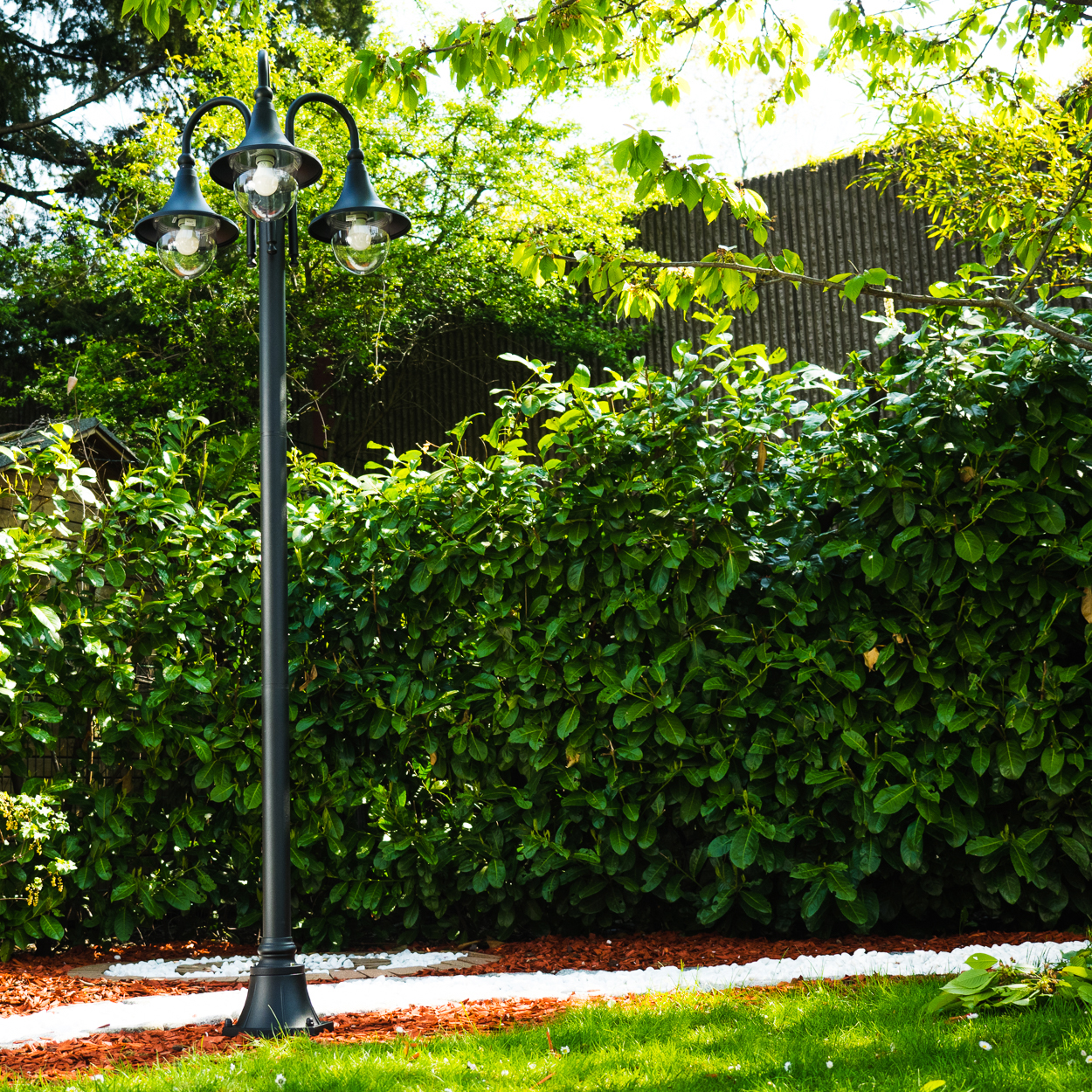 Eine schwarze Laterne mit drei Lampen steht vor einer grünen Hecke. Ein weißer Weg und Gras sind im Vordergrund.