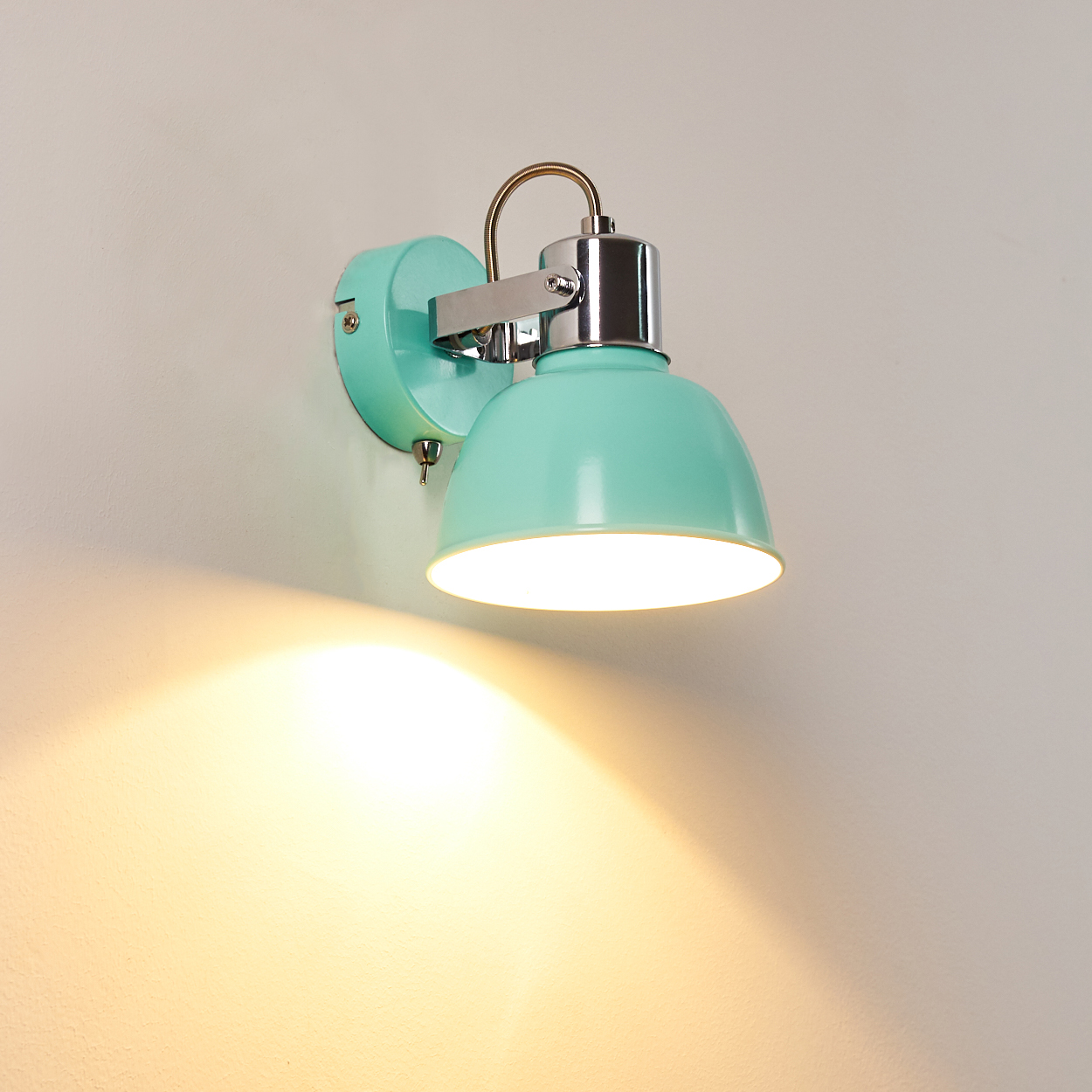 Eine mintgrüne Wandleuchte ist an einer weißen Wand montiert. Die Lampe ist an und wirft ein warmes Licht.
