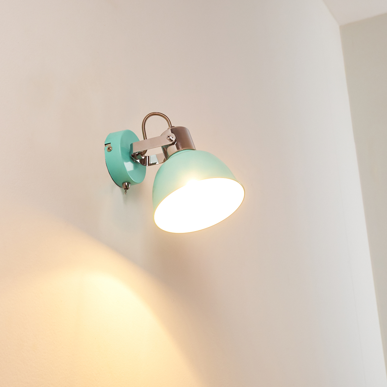 Eine mintgrüne Wandleuchte ist an einer weißen Wand befestigt und strahlt helles Licht aus. Die Lampe hat einen Metallarm und -schirm.