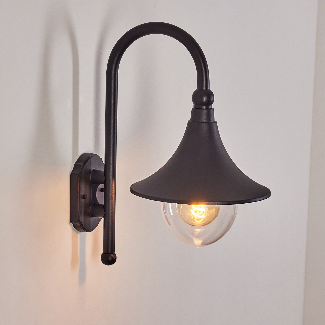 Eine schwarze Außenwandleuchte. Die Lampe hat einen gebogenen Arm, einen konischen Schirm und einen durchsichtigen Glaskolben mit einer Glühbirne.