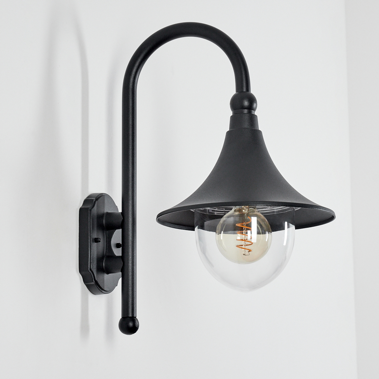 Eine schwarze Wandleuchte mit gebogenem Arm und einem klaren Glaskolben. Der Hintergrund ist eine schlichte weiße Wand.