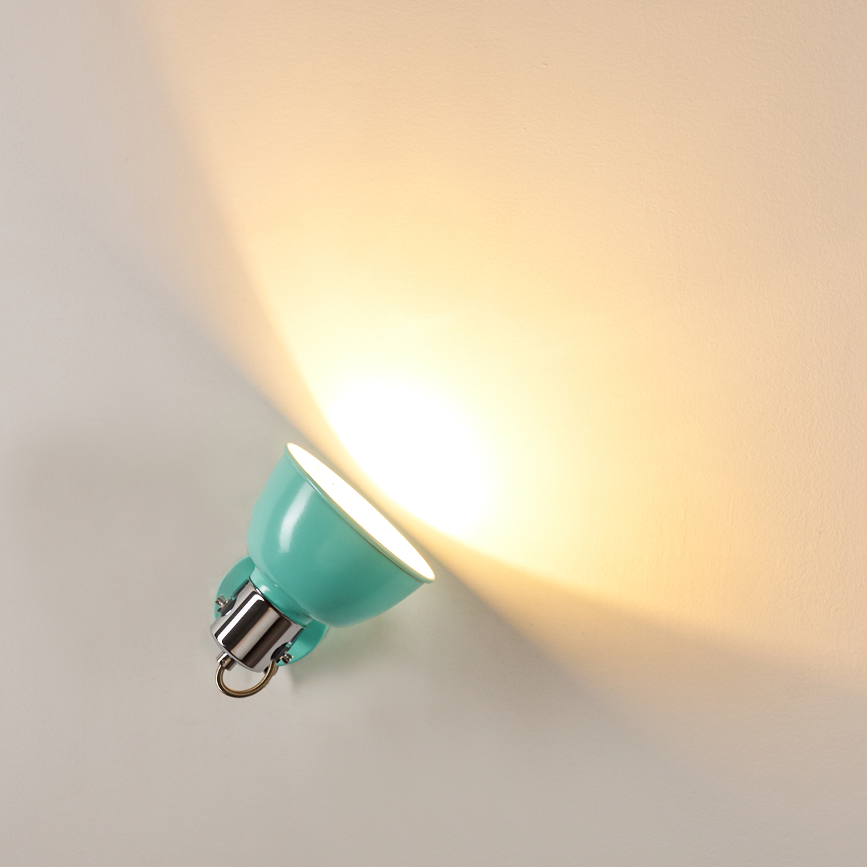 Eine türkisfarbene Wandleuchte mit silbernem Sockel strahlt Licht auf eine weiße Wand. Das Licht ist hell und gelb.