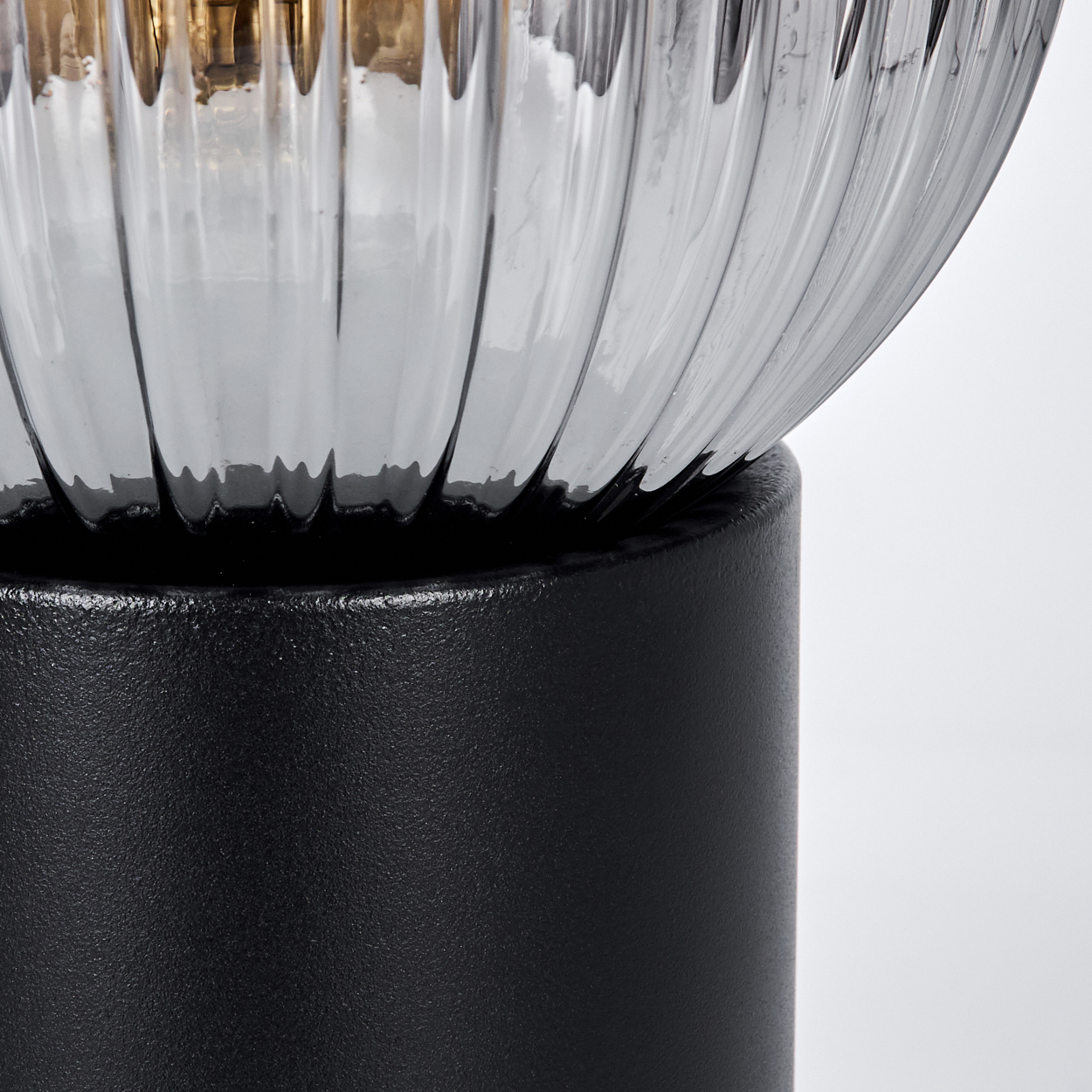 Nahaufnahme einer Lampe. Die Lampe hat einen schwarzen Sockel und einen klaren, gerippten Glasschirm. Der Hintergrund ist weiß.