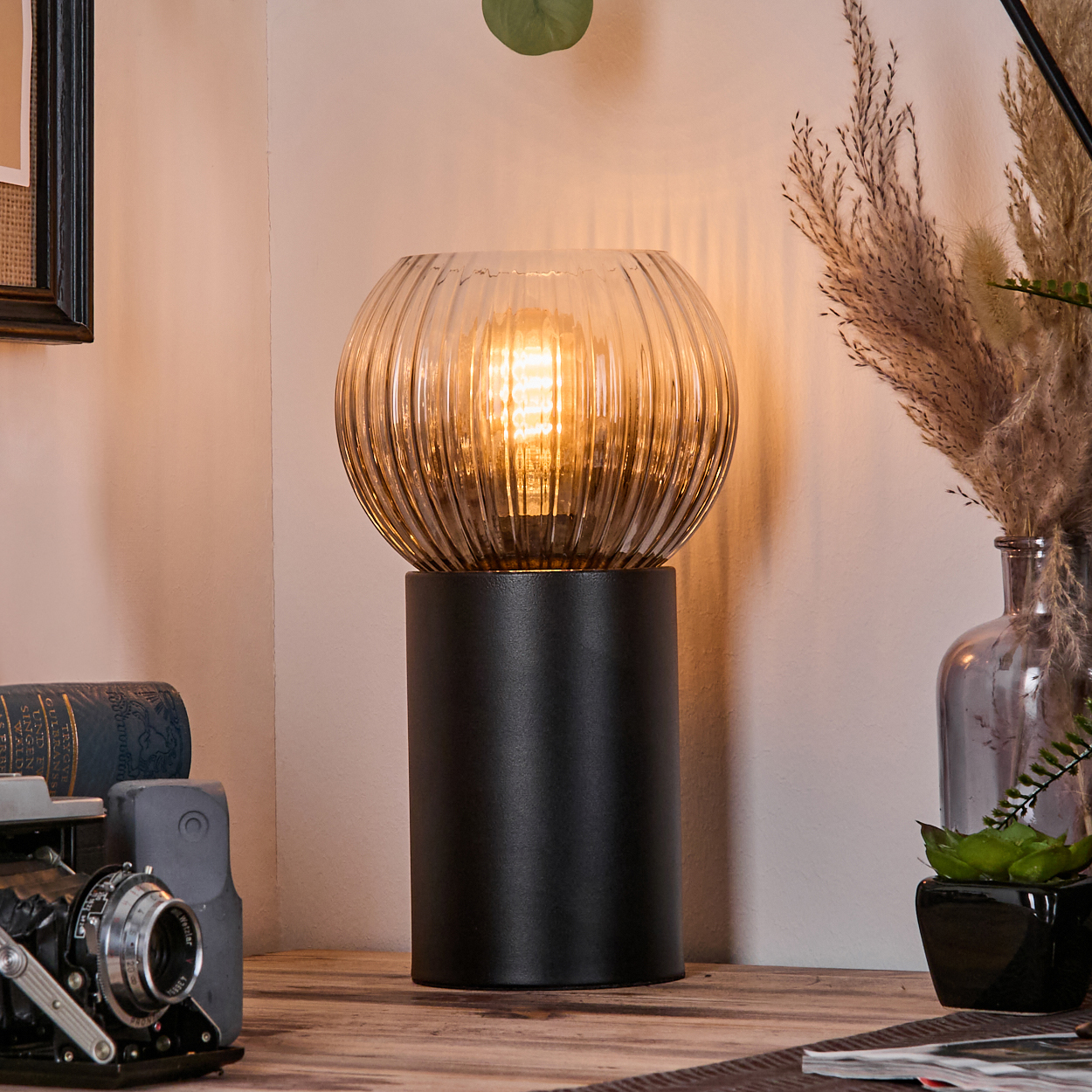 Eine beleuchtete Lampe auf einem Tisch mit Kamera und Büchern. Rechts eine Vase mit Pampasgras. Warmes Licht und gemütliche Atmosphäre.