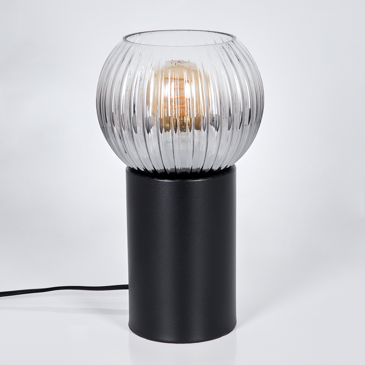 Eine moderne Lampe mit gerippter Glaskugel und schwarzem zylindrischen Sockel, beleuchtet. Ein schwarzes Kabel.