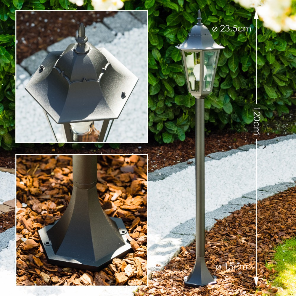Dunkle Metall-Gartenlaterne. Sie hat oben eine Laterne und steht auf einem Sockel. Der Hintergrund umfasst einen Garten mit Pflanzen.