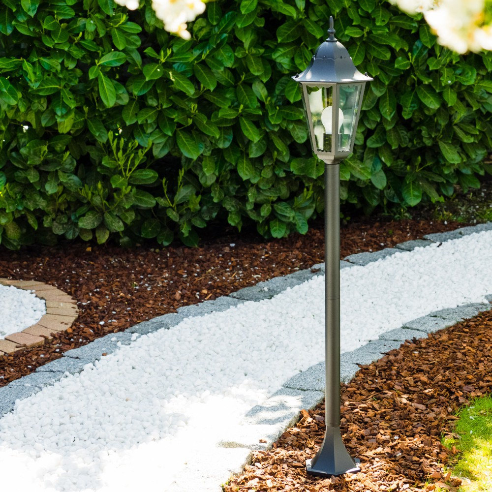 Eine Gartenszene mit einer Laterne, einem weißen Steinweg, einem grünen Busch und braunem Mulch. Helles Sonnenlicht beleuchtet die Szene.
