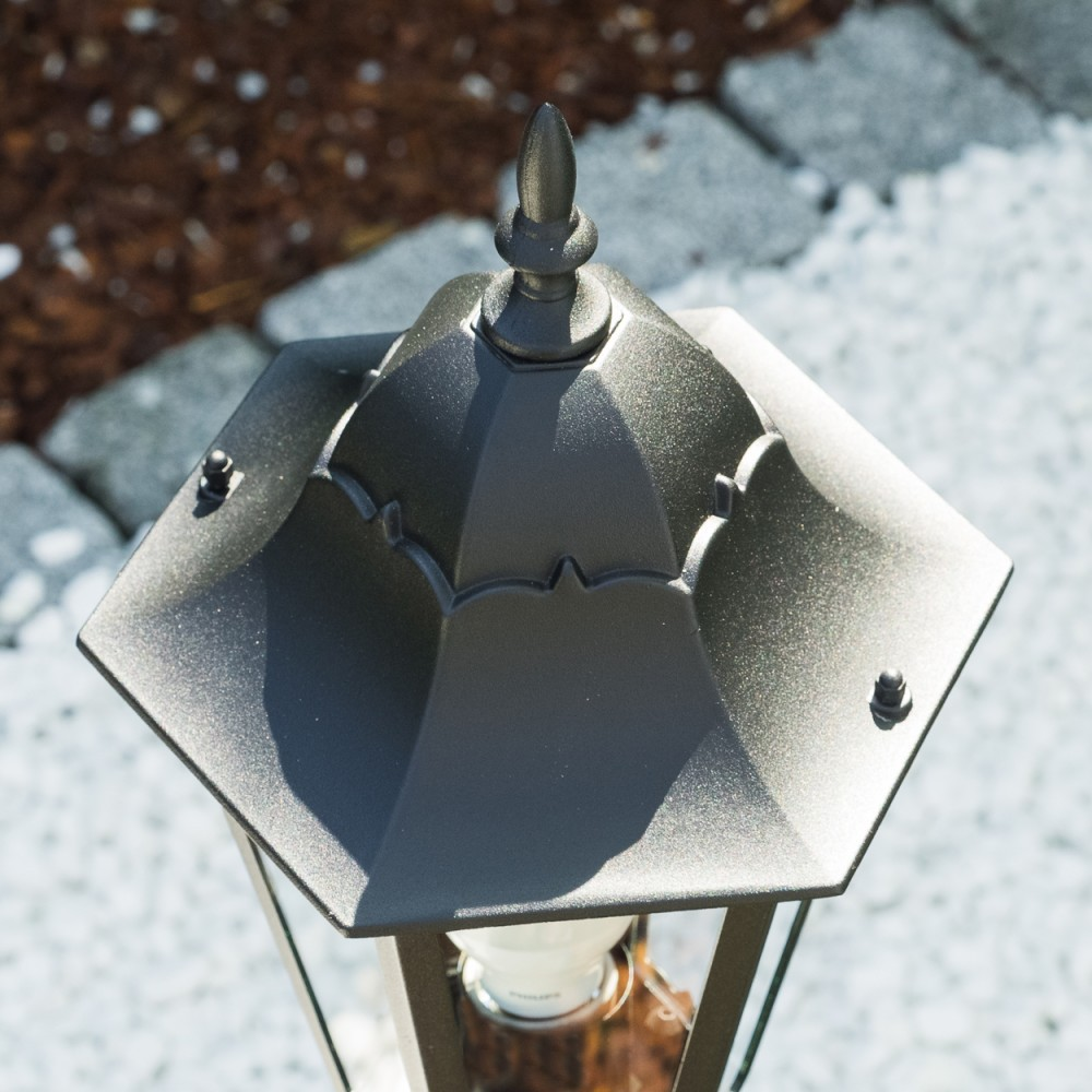 Nahaufnahme einer schwarzen, sechseckigen Außenlampe. Die Lampe hat eine dekorative Oberseite und steht vor einem Hintergrund aus Steinen und Holzspänen.