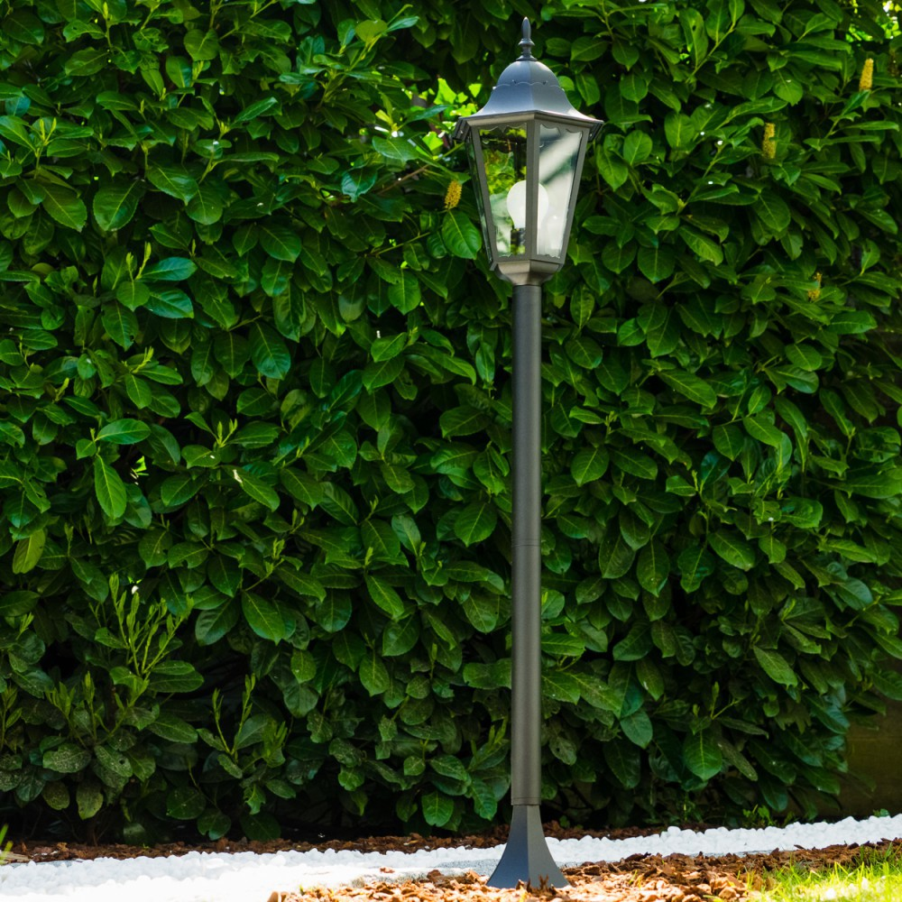 Eine schwarze Laterne steht vor einer üppigen grünen Hecke. Weiße Steine und brauner Mulch befinden sich am Fuß der Hecke und der Lampe.