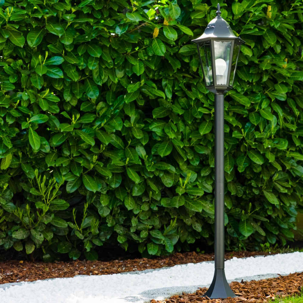 Eine schwarze Laterne steht vor einer grünen Hecke. Weiße Steine und brauner Mulch säumen den Weg.