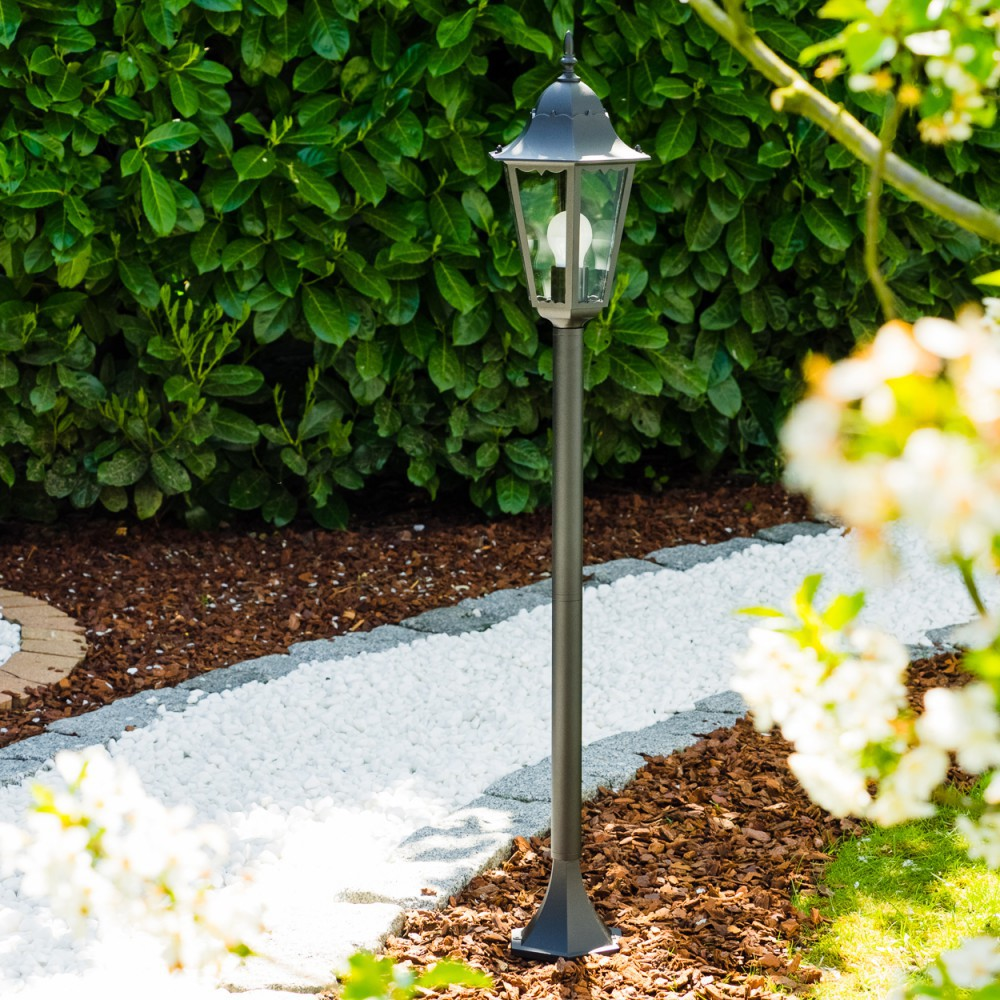 Dunkle Metall-Laterne im Garten. Grüner Buschhintergrund. Weißer Steinweg und brauner Mulchvordergrund mit weißen Blumen.