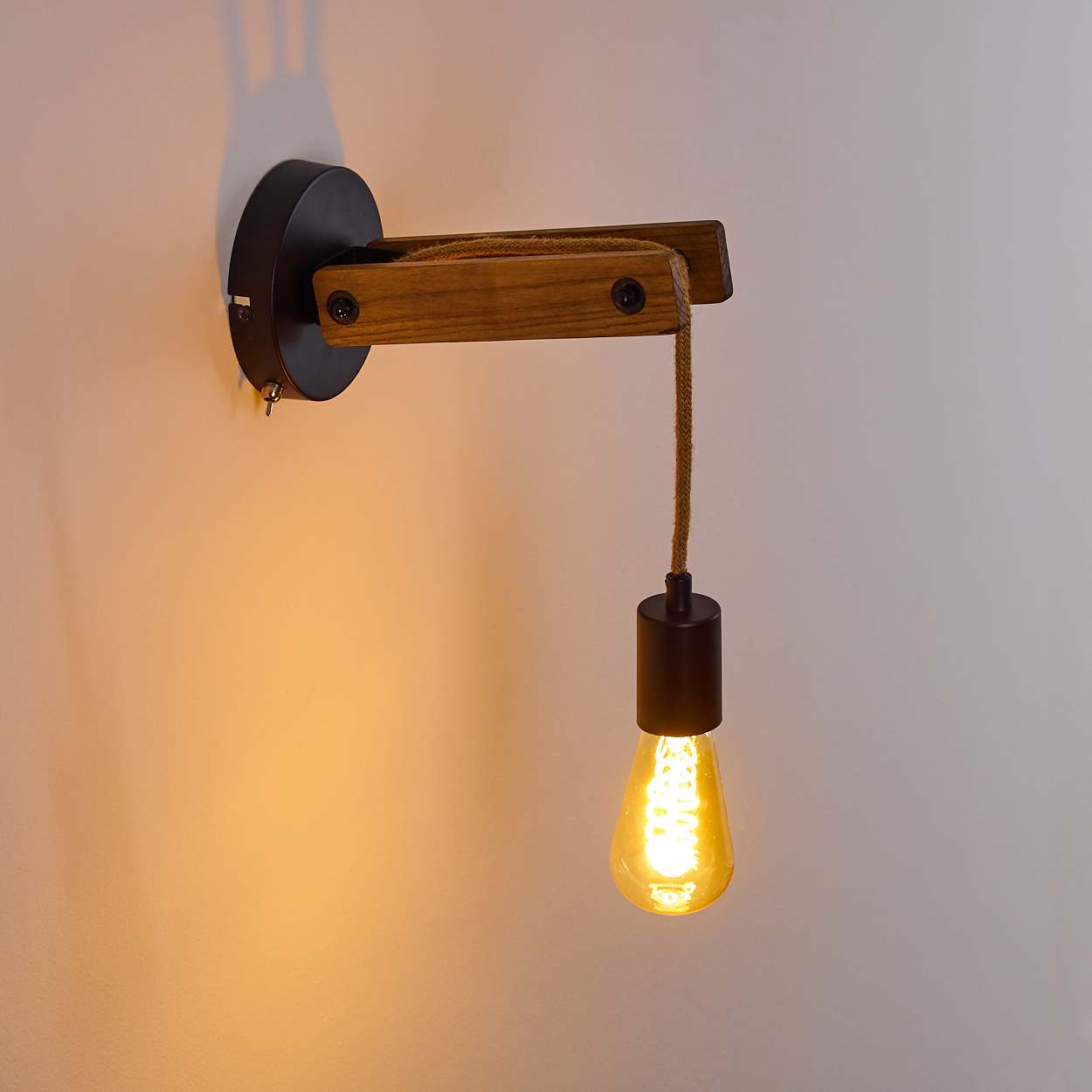 Eine an der Wand montierte Holzlampe mit einer hängenden Glühbirne leuchtet vor einer hellen Wand. Die Lampe hat einen schwarzen Sockel und ein braunes Seil.