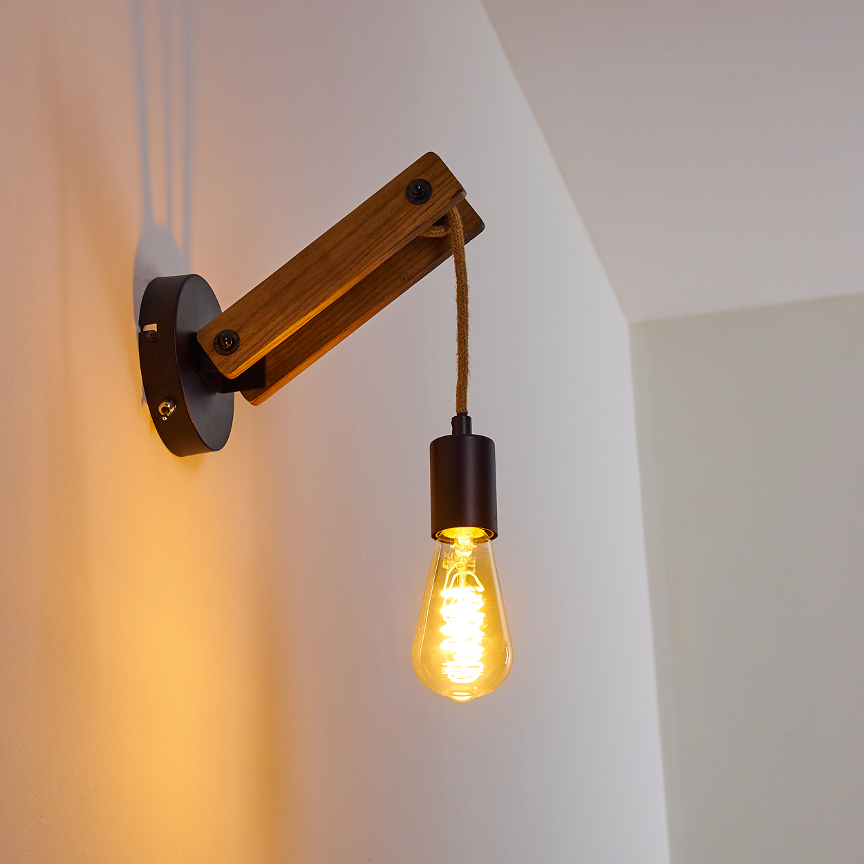 Eine Wandlampe aus Holz mit einer hängenden Glühbirne. Die Lampe hat einen braunen Holzarm und einen schwarzen Sockel, montiert an einer weißen Wand.