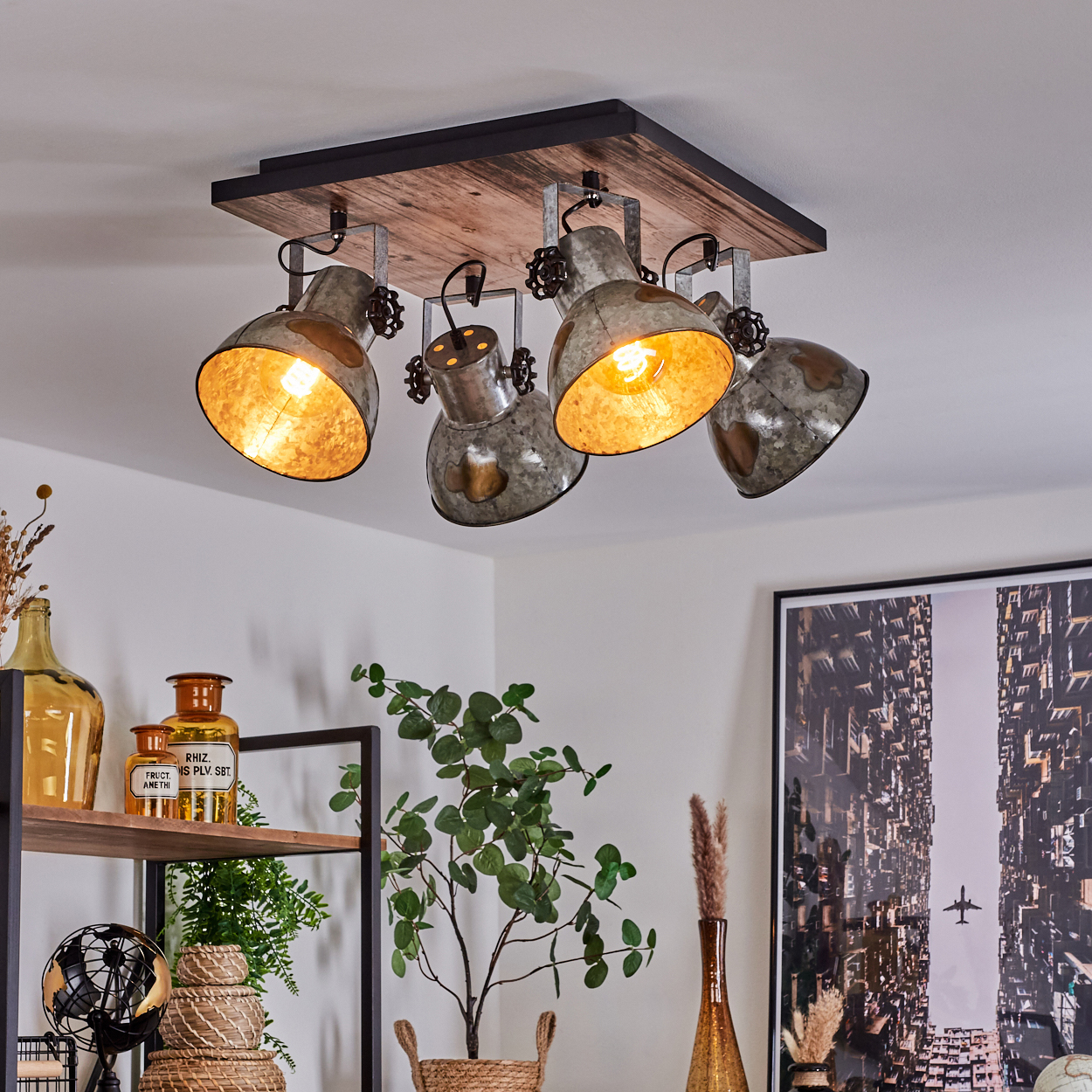 Deckenleuchte mit vier Strahlern, Holzsockel, Metallschirme. Innenraum mit Pflanzen, Regalen und einem Stadtbild an der Wand.