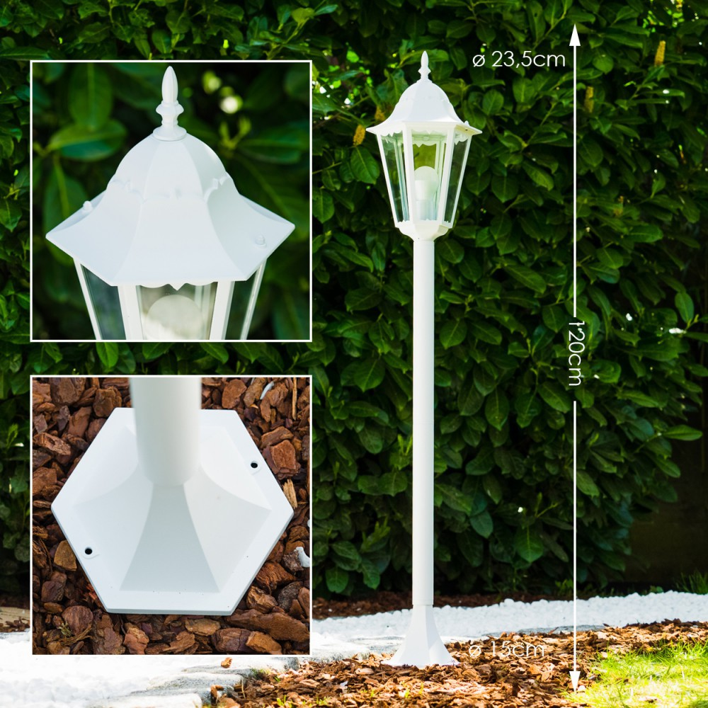 Eine weiße Außenlampe. Sie hat einen sechseckigen Sockel, eine lange Stange und eine dekorative Oberseite mit Glasplatten. Hintergrund ist grünes Laub.