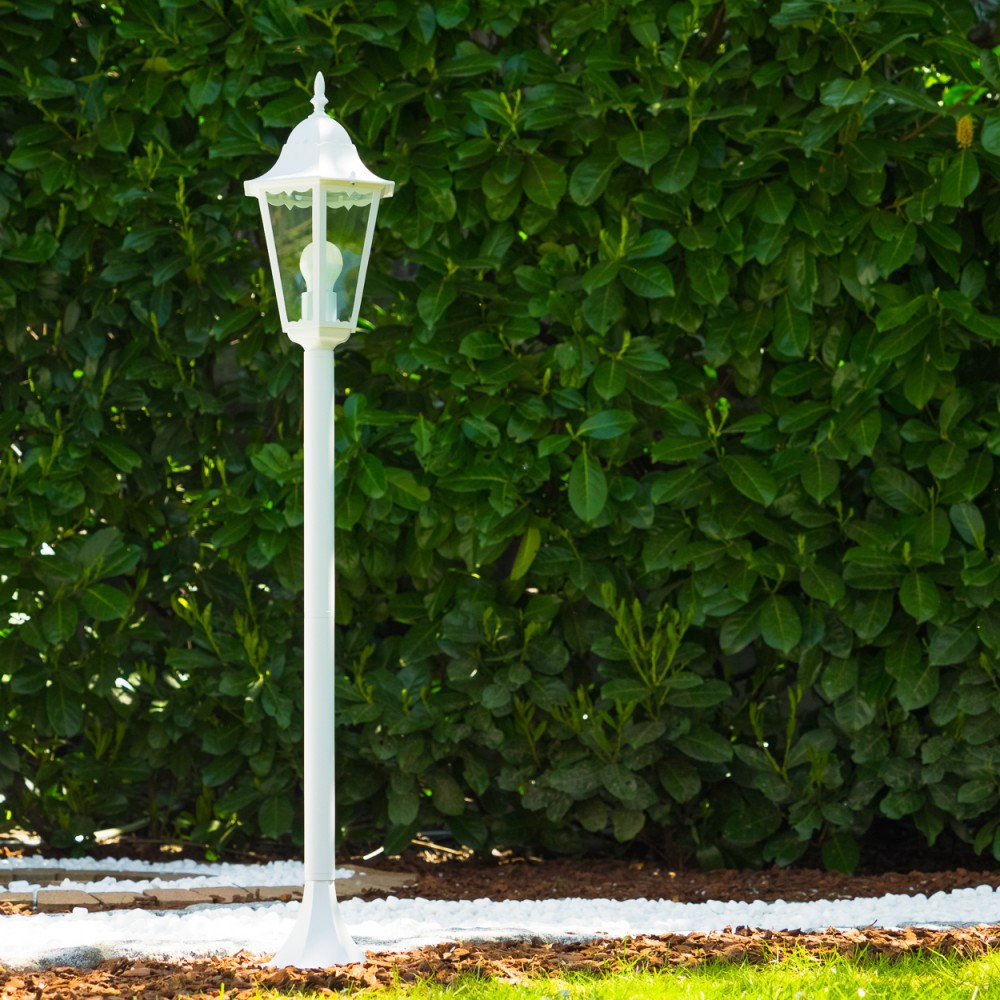 Eine weiße Außenlampe steht vor einer grünen Hecke. Die Lampe steht auf einem Pfahl und hat ein Glasgehäuse für das Licht.