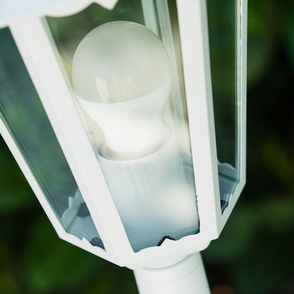 Nahaufnahme einer weißen Außenlampe mit Glühbirne im Inneren, umgeben von Glasscheiben. Grünes Laub im Hintergrund.