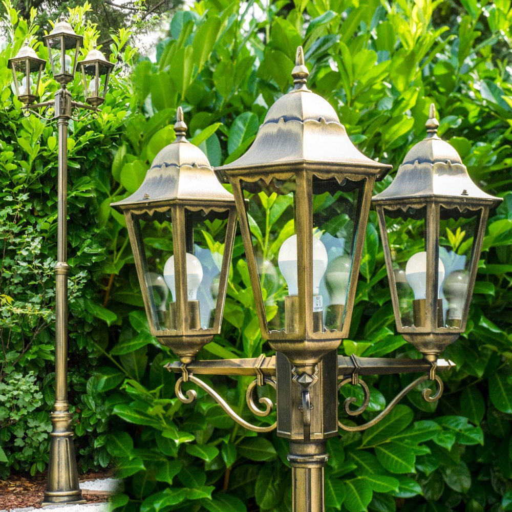 Eine bronzefarbene Laterne mit drei Lampen, jede mit einer Glühbirne. Grünes Laub bildet den Hintergrund.