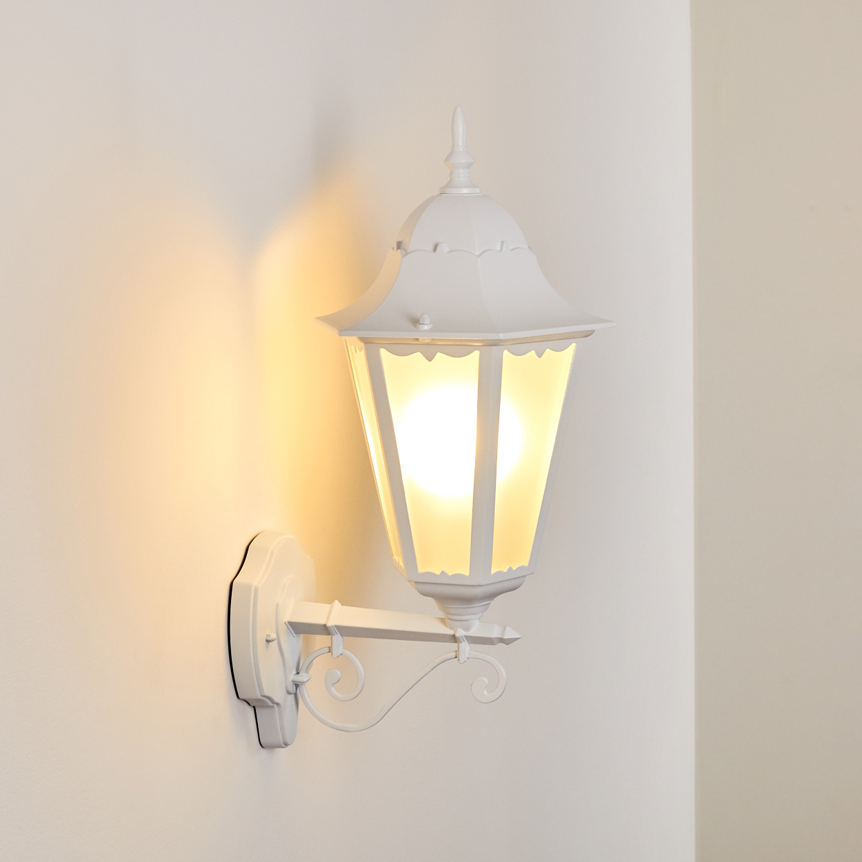 Eine weiße, verzierte Wandlampe ist an einer weißen Wand befestigt. Die Lampe hat ein beleuchtetes, gelb getöntes Glasgehäuse.