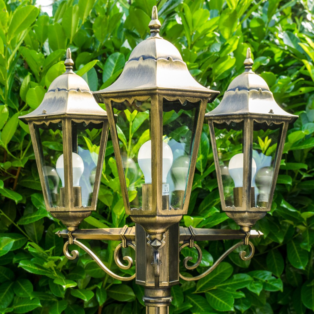 Antike Straßenlaterne mit drei Lampen und weißen Glühbirnen vor grünem Hintergrund.
