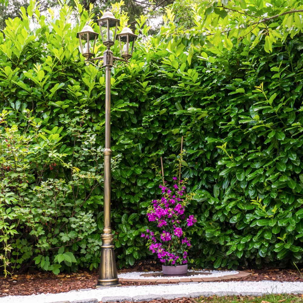 Eine bronzefarbene Laterne steht vor einer grünen Hecke. Rechts befinden sich lila Blumen in einem Topf. Weiße Steine und Mulch am Boden.
