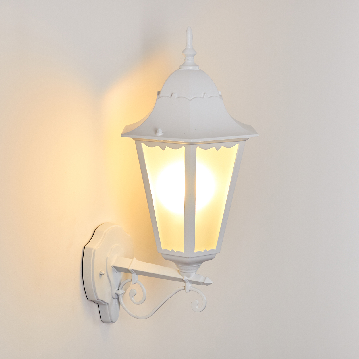 Eine weiße Außenlaterne ist an einer Wand befestigt. Sie hat ein dekoratives Design und wird von warmem Licht beleuchtet.
