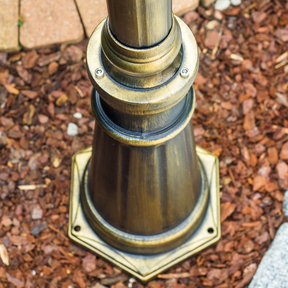Nahaufnahme eines bronzenen Lampenfußes auf braunem Mulch. Die Basis hat eine sechseckige Form. Der Hintergrund zeigt Ziegel und Stein.