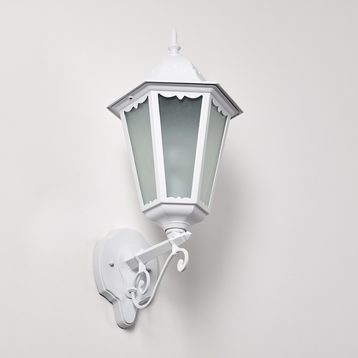 Eine weiße, verzierte Außenlaterne ist an einer weißen Wand befestigt. Die Laterne hat eine sechseckige Form mit Milchglasscheiben.