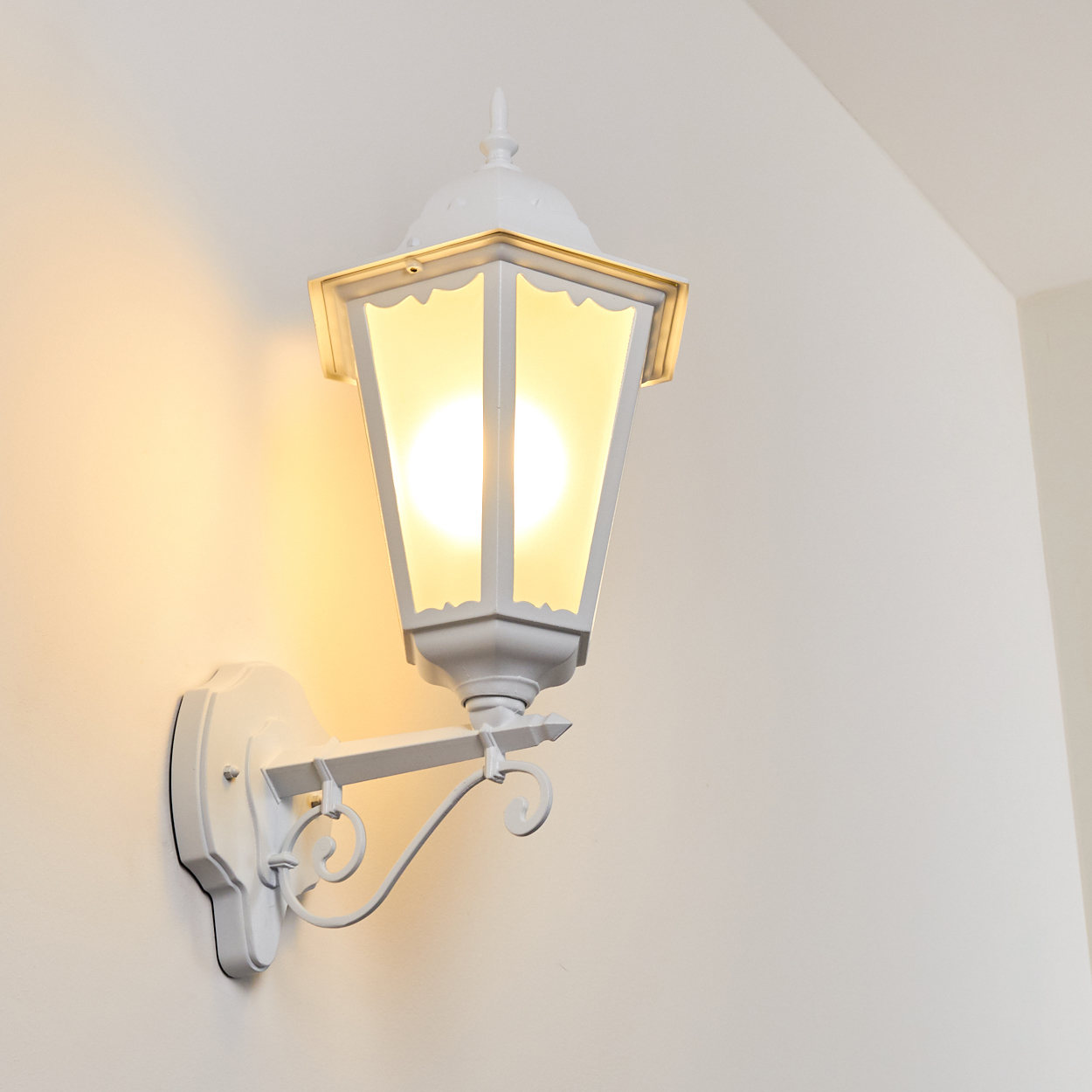 Weiße Wandlampe mit warmem Licht. Die Lampe ist an der Wand befestigt. Die Wand ist weiß.