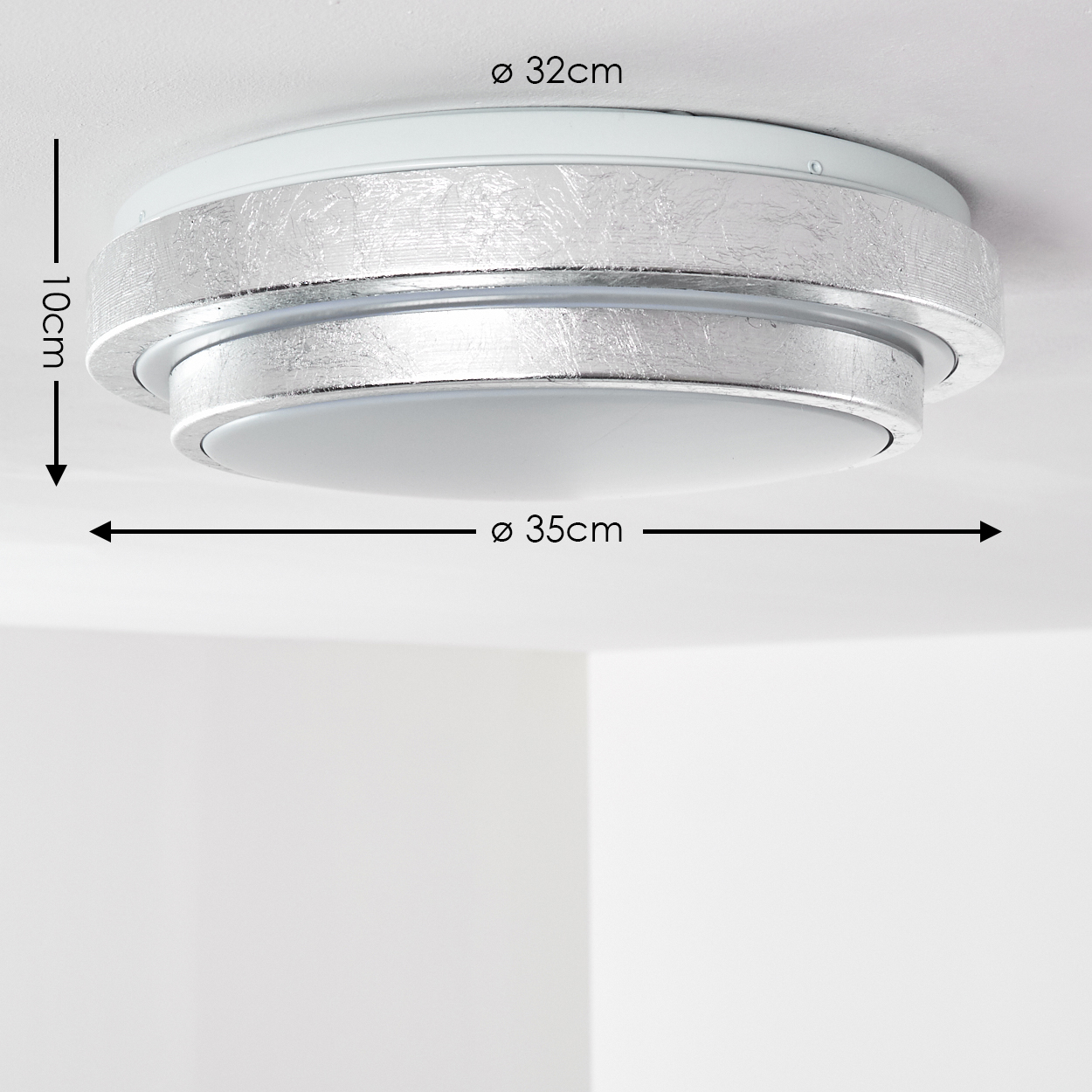 Eine Deckenleuchte mit silbernem Metallband, weißer Basis und beschrifteten Abmessungen. Das Licht ist an einer weißen Decke.