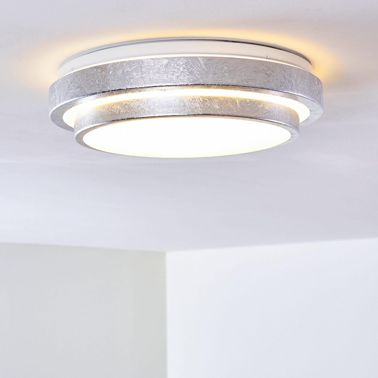 Eine moderne Deckenleuchte in Silber und Weiß. Das Licht ist an und beleuchtet die weiße Decke und eine Ecke der weißen Wände.